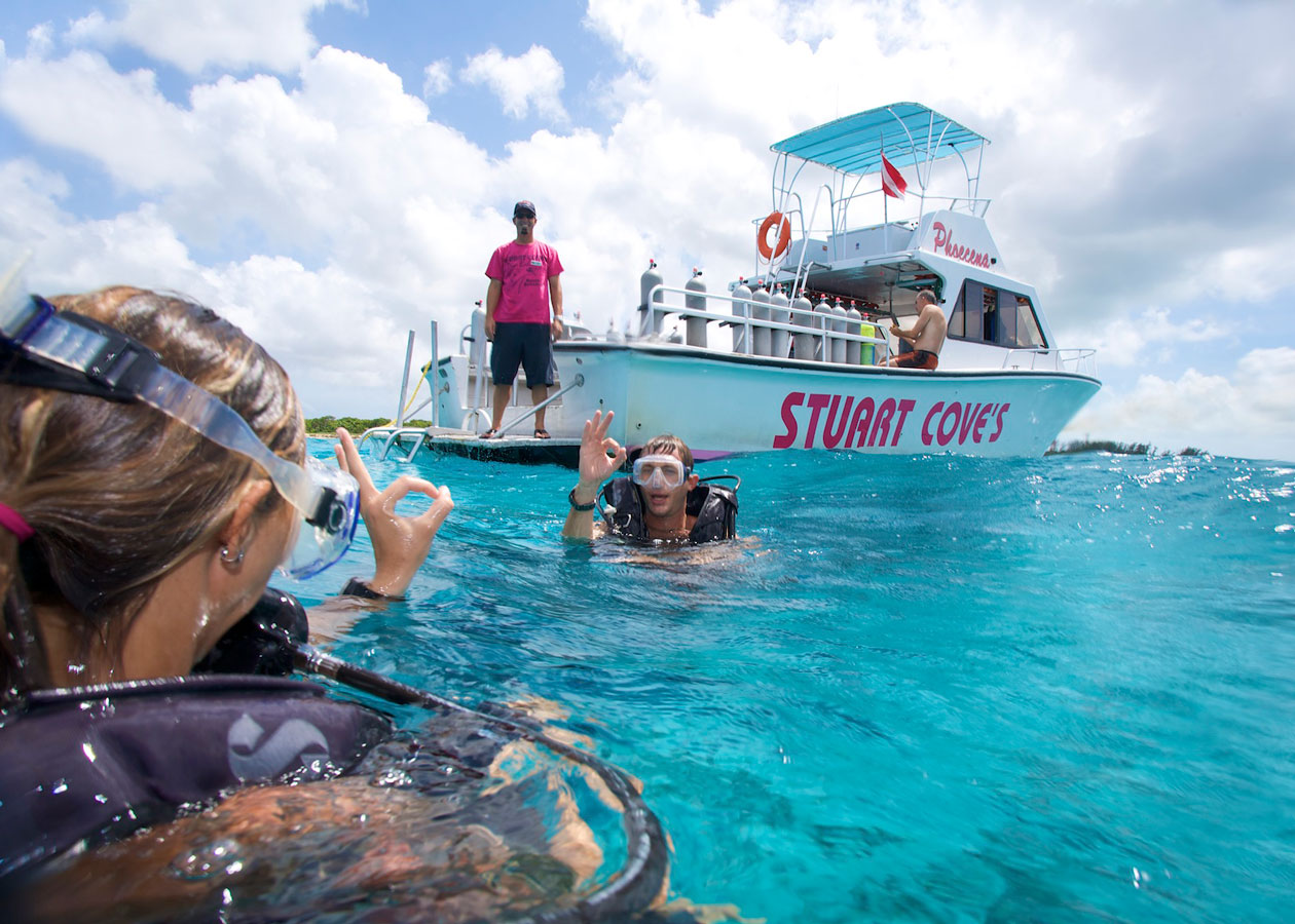 two scuba divers giving the ok signal in the blue waters of the bahamas with the boat names stuart cones in the background with crew members on it